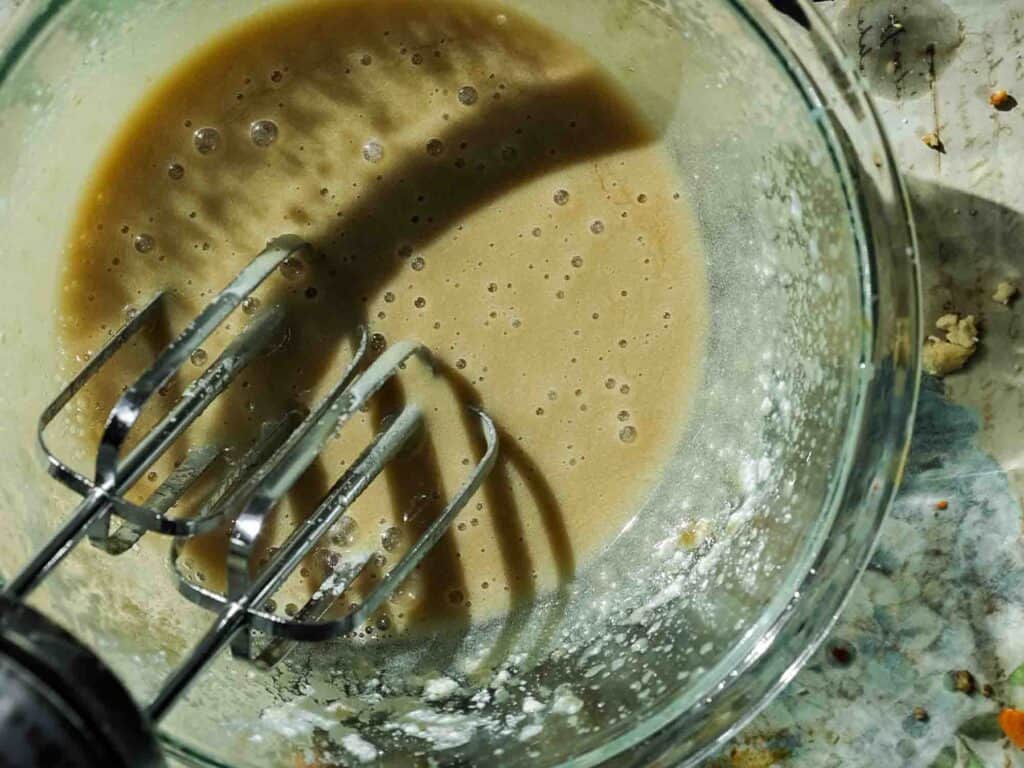 A glass bowl containing tan batter is shown from above, partially mixed with an electric mixer with two beaters. The bowl rests on a speckled surface, and light casts a shadow of the beaters onto the batter.